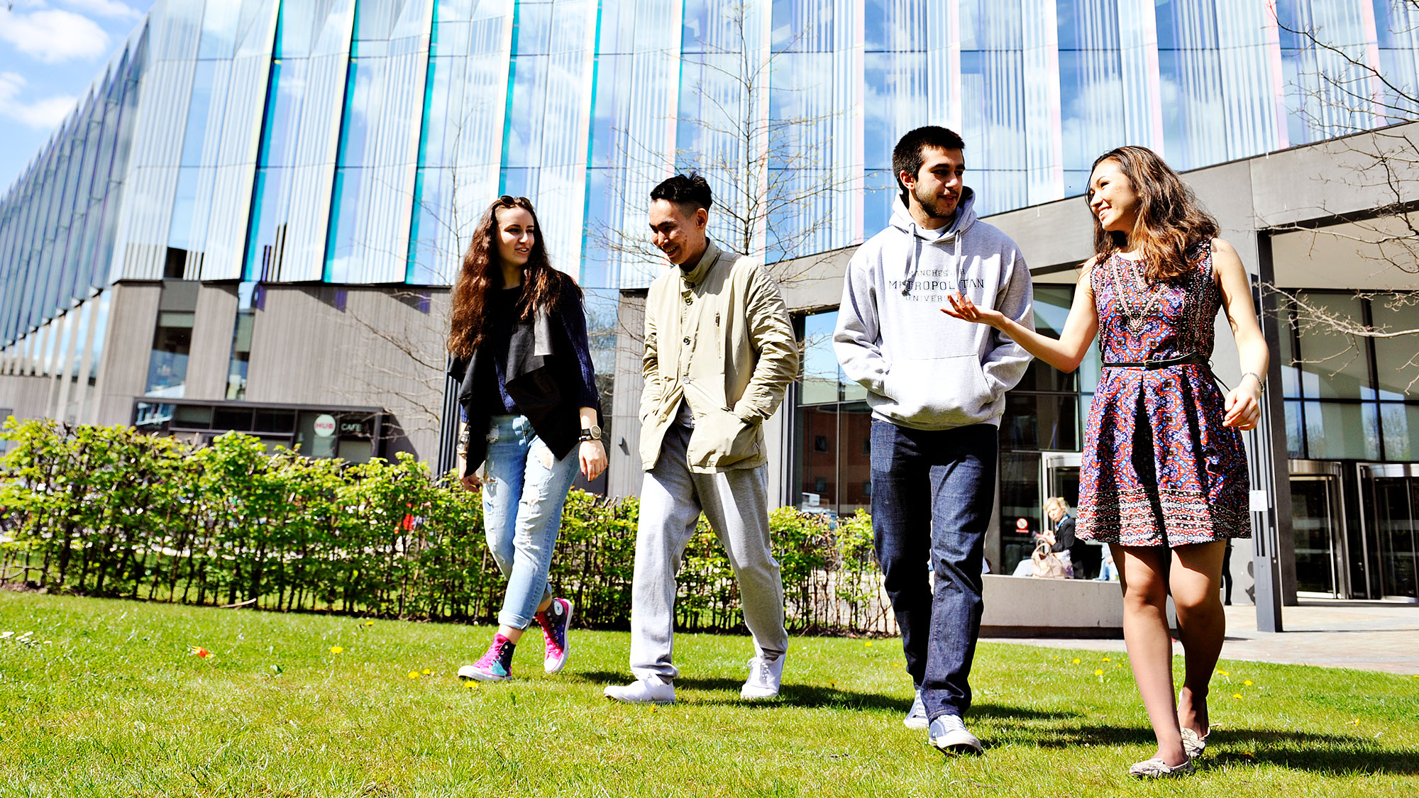 Four international students from INTO Manchester walk on lawn in front of Manchester Metropolitan University building. 