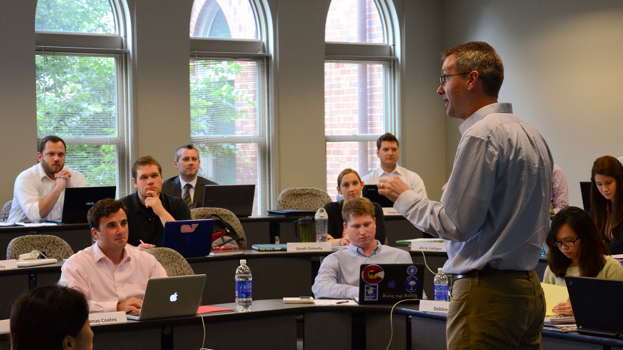 Saint Louis University students sit at rows of desks with laptops and listen to professor’s lecture in classroom.
