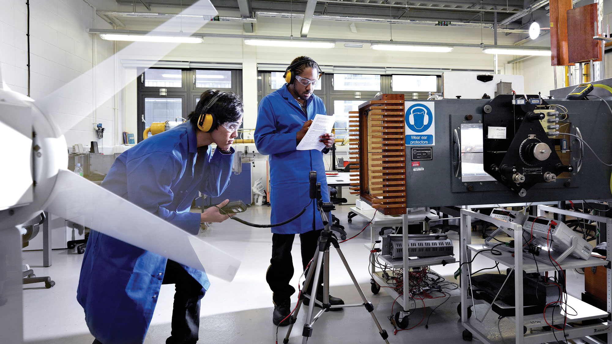 Two Manchester Metropolitan University students conduct experiment wearing sound-proof ear muffs, goggles, and blue coats in physics laboratory. 