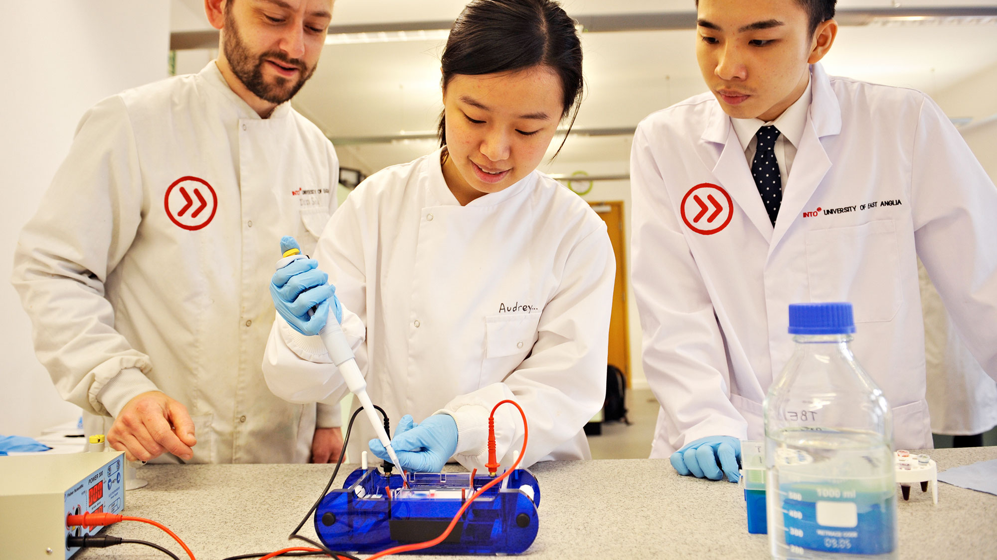 INTO University of East Anglia international students conduct experiment involving electronic wires, wear latex gloves and INTO-branded white coats. 