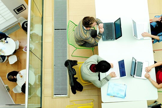 Overhead view of two students sitting on green and yellow chairs at desk and working on laptops in INTO University of East Anglia Centre. 