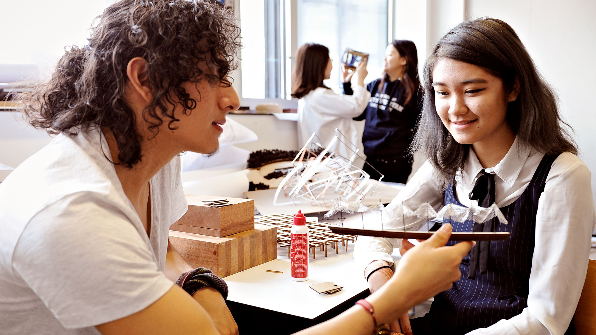 INTO Manchester international students look at art object made of wood, small posts, and airy fabric, while sitting at desk holding glue and wood.